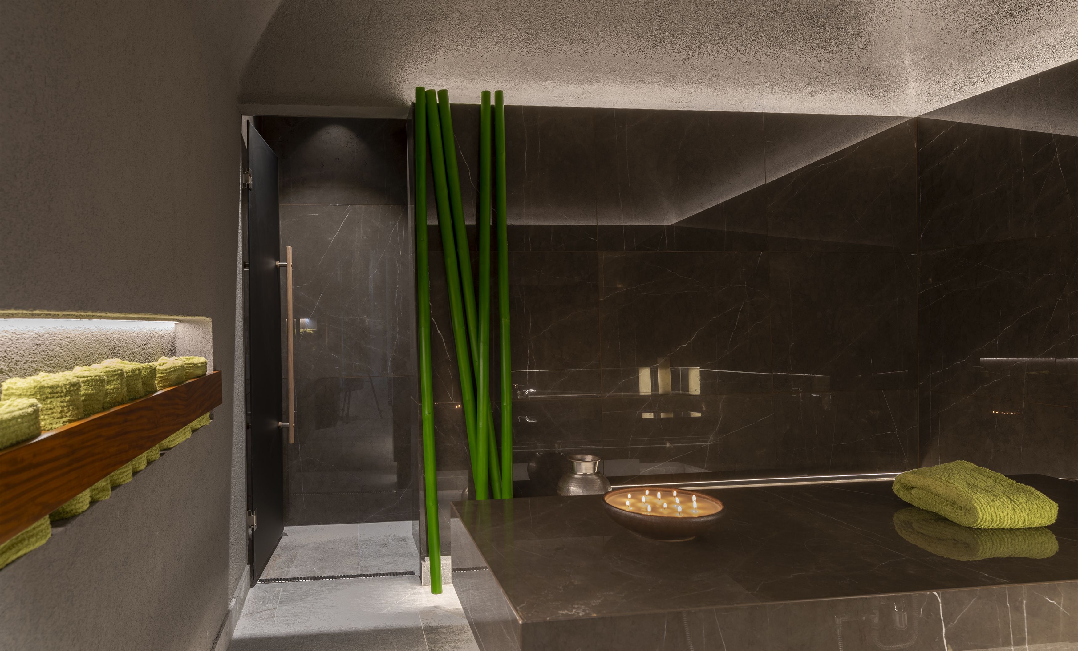 A modern bathroom with a dark marble wall, a wooden shelf with green towels, a green towel on the floor, and a small bowl with candles.