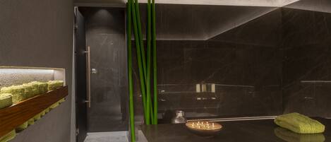 A modern bathroom with a dark marble wall, a wooden shelf with green towels, a green towel on the floor, and a small bowl with candles.