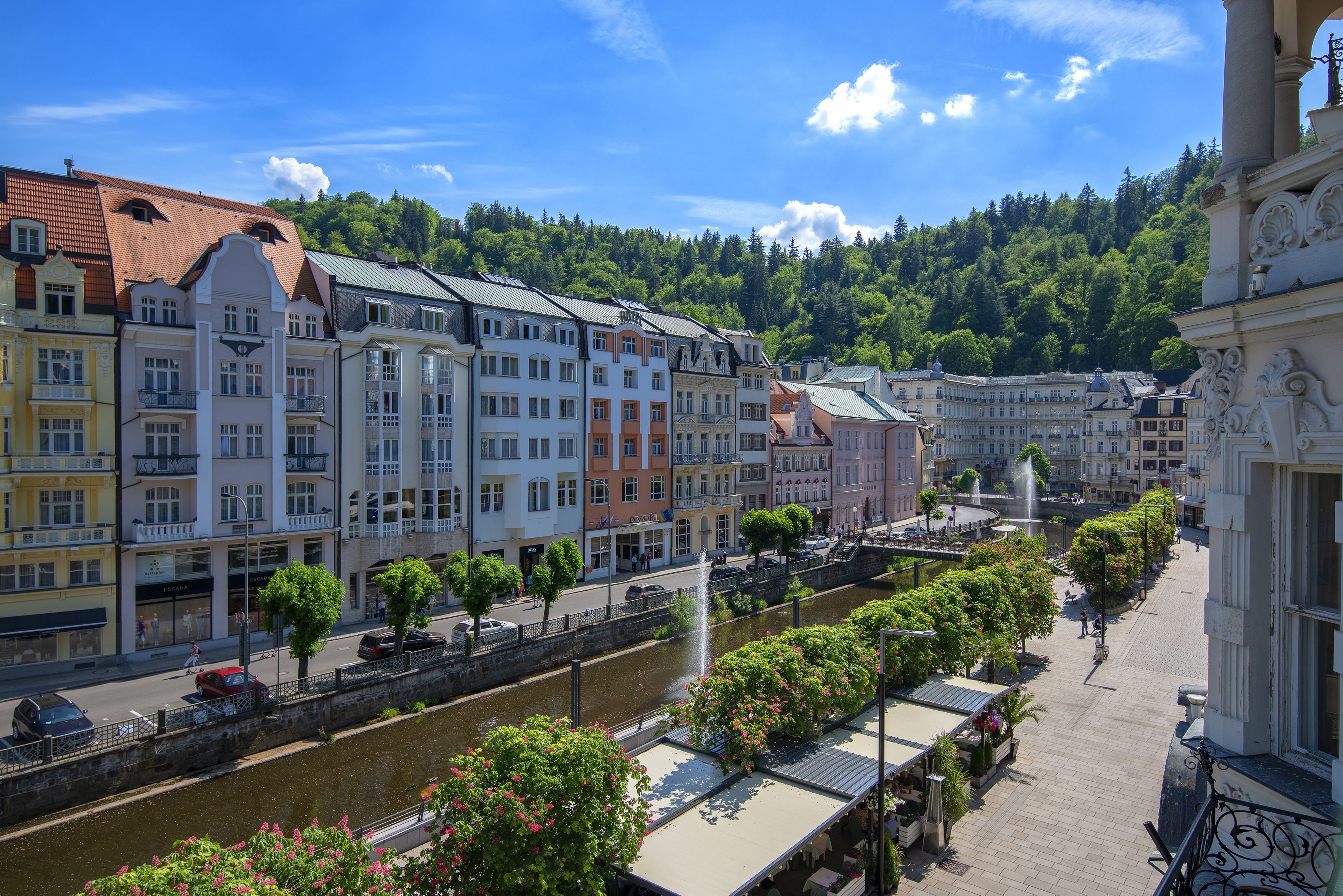 A riverside promenade with buildings, a fountain, and a bridge.