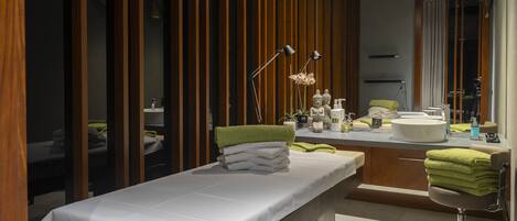A modern spa room with a massage table, a small seating area, and a vanity with a mirror.