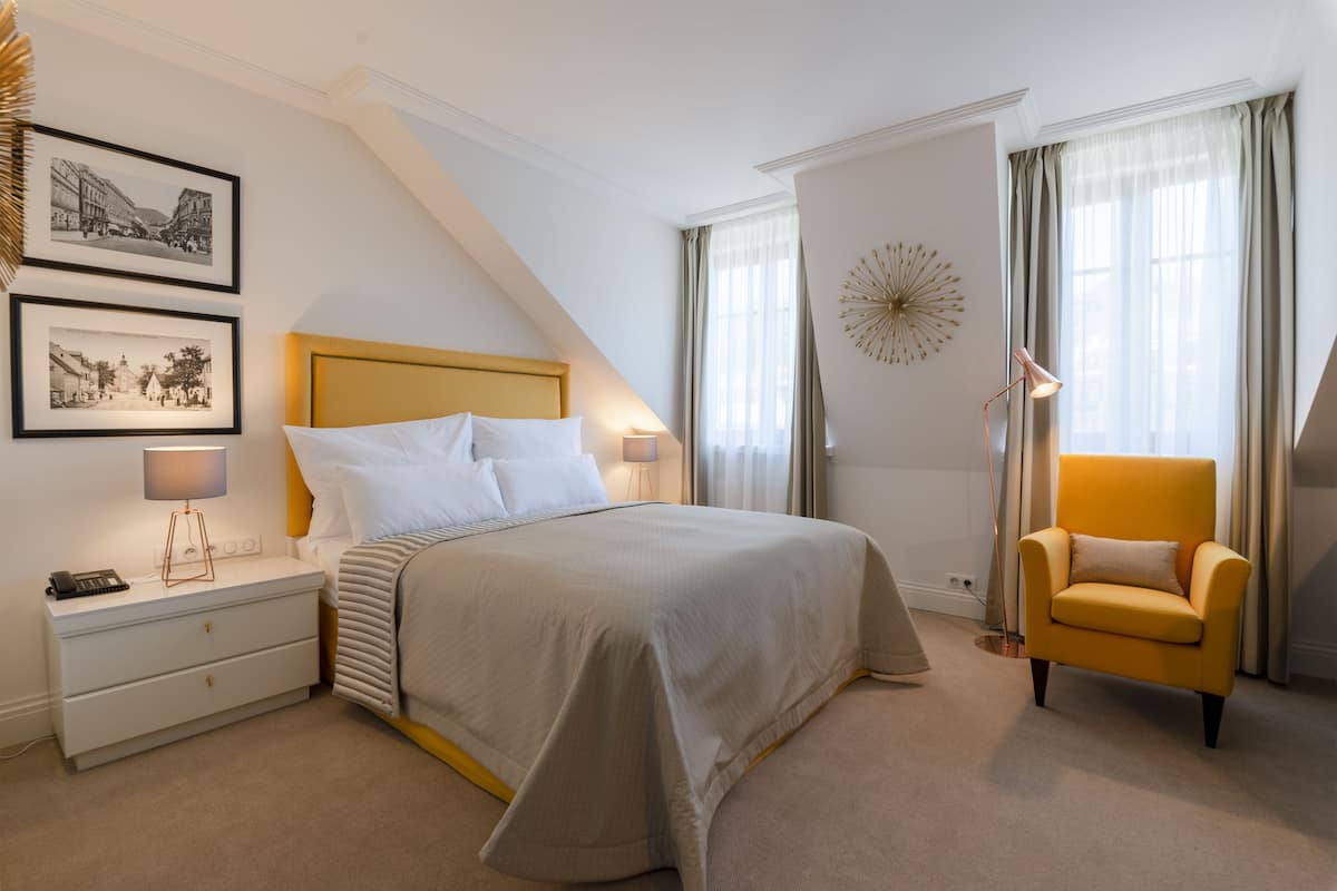Een slaapkamer met een bed, nachtkastje, lamp, stoel en twee foto’s in lijsten aan de muur.