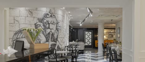 A modern hotel lobby with a marble floor, black and white striped pattern, and a large wall mural. There are dining tables and chairs arranged for guests.