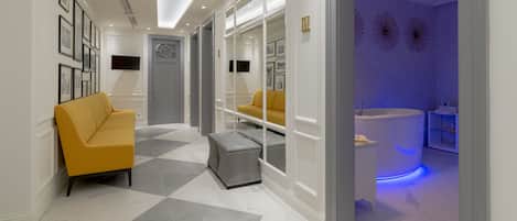 A modern hotel lobby with a yellow sofa, a checkered ottoman, and a reception desk with a blue-lit sink.