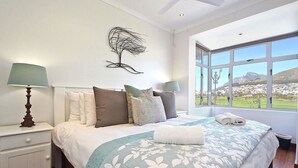 Ein Schlafzimmer mit einem Bett, zwei Nachttischlampen, einem Wandbild und einem Fenster mit Blick auf eine Landschaft.
