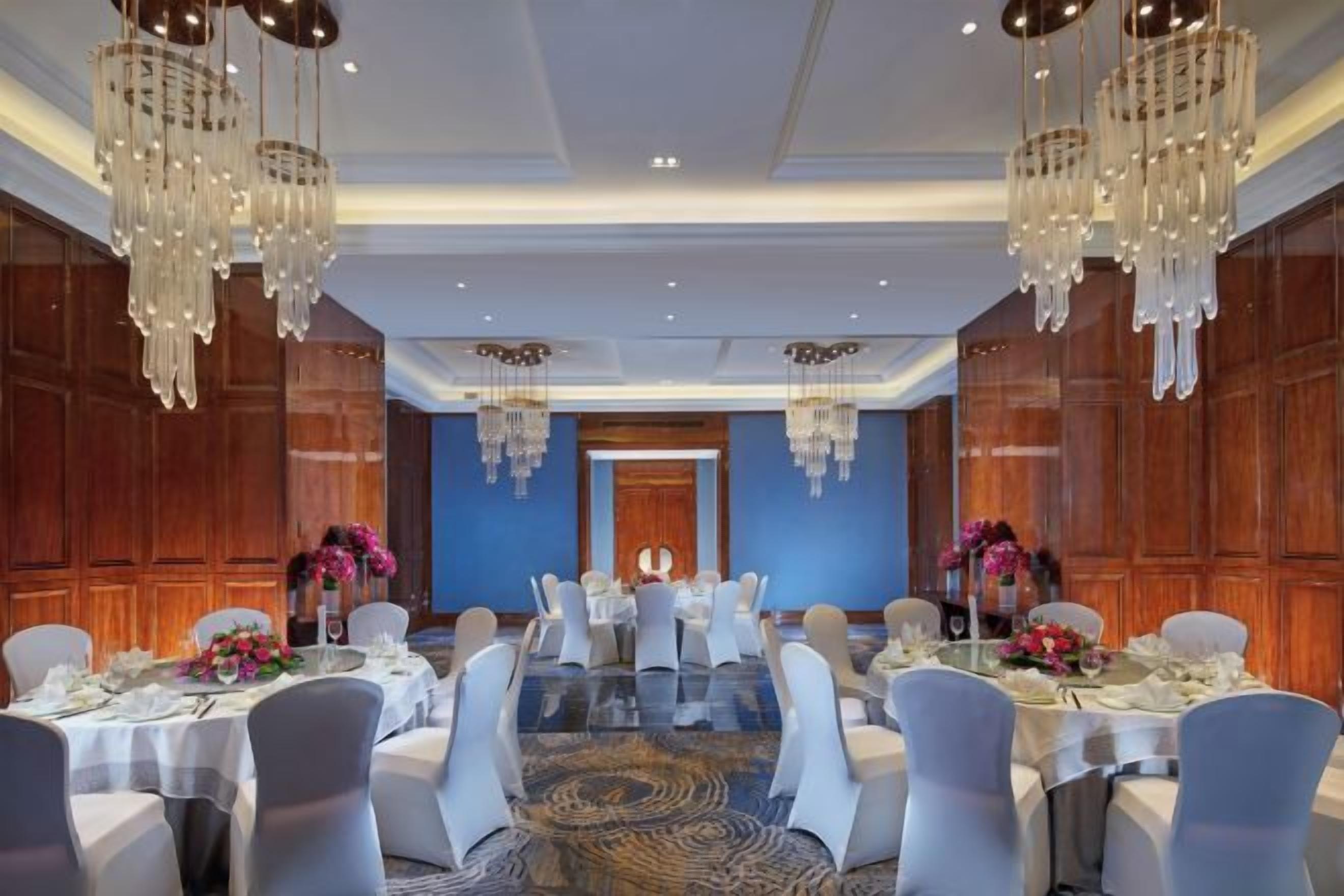 A banquet hall with round tables, white chairs, chandeliers, and floral centerpieces.