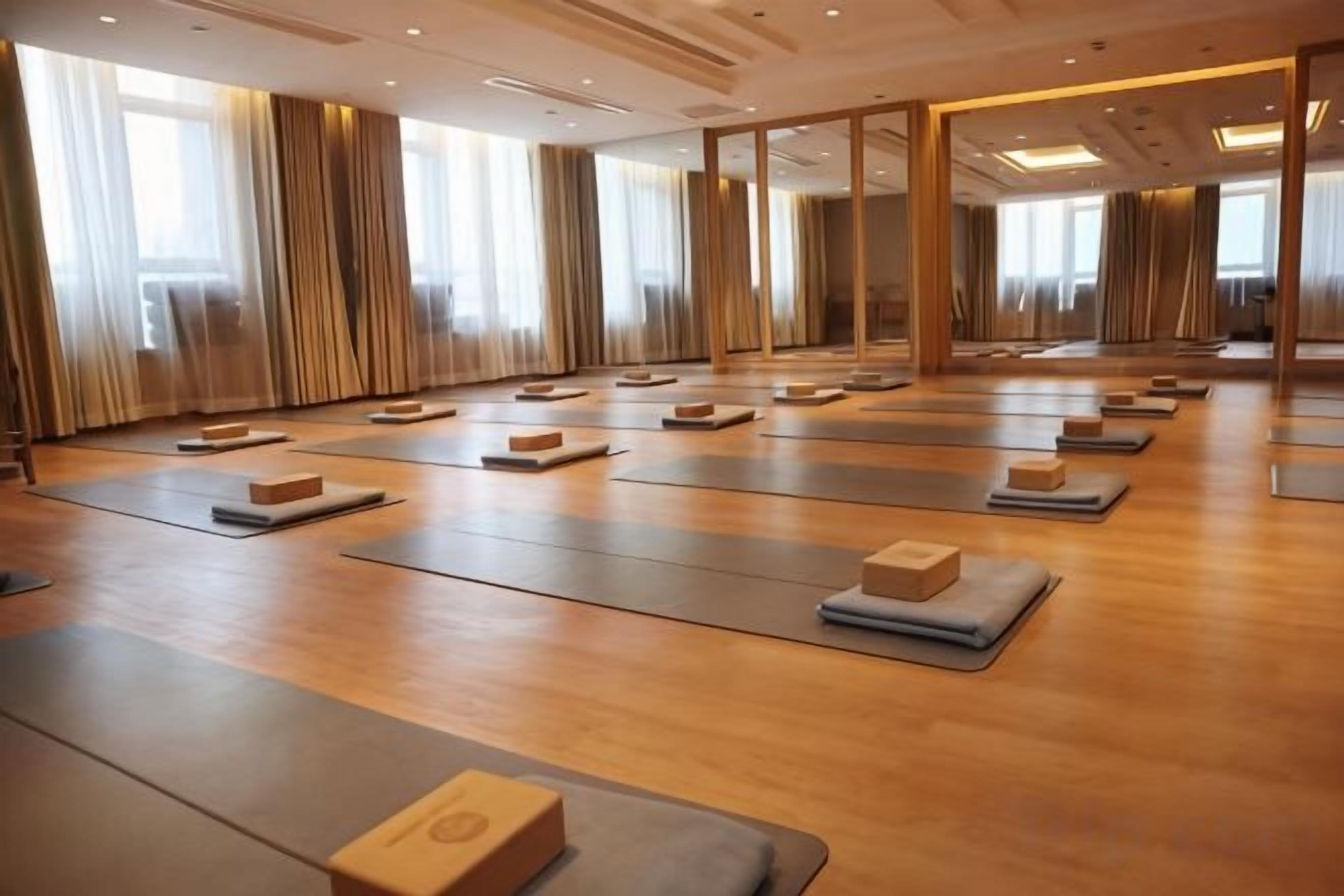 A spacious room with multiple yoga mats arranged in rows, large windows with curtains, and a wooden floor.