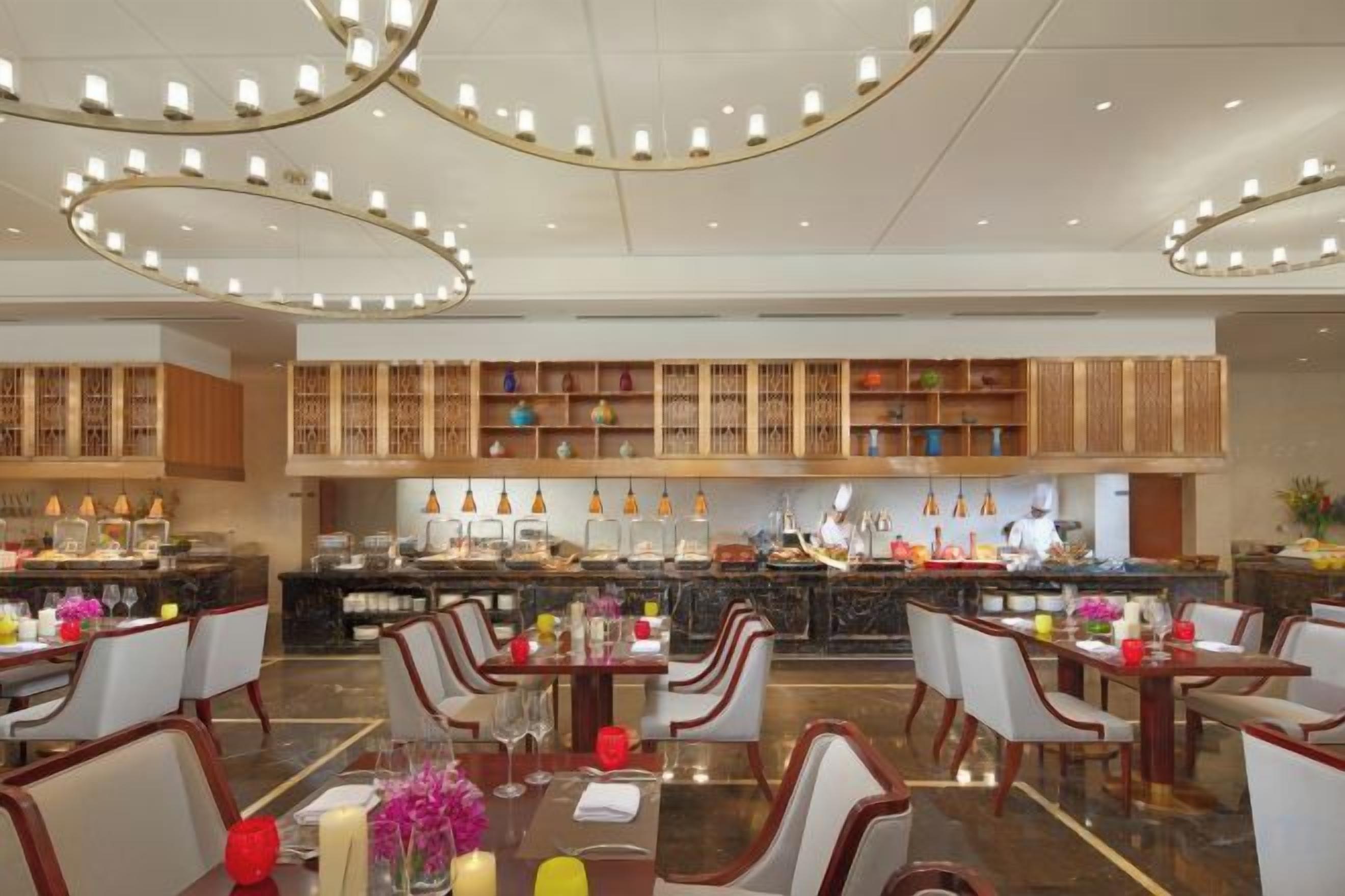 A spacious hotel dining area with multiple tables and chairs, a central bar area, and a kitchen in the background.