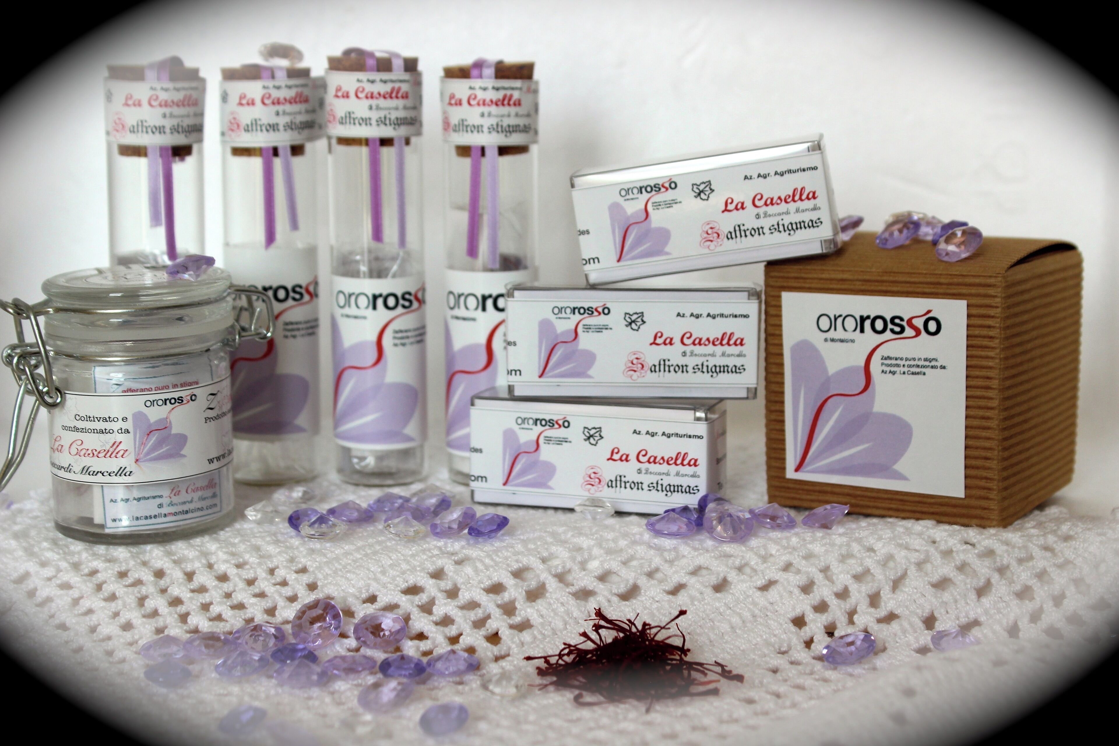 Uma seleção de produtos de açafrão La Casella, incluindo potes, caixas e uma caixa de madeira, dispostos sobre uma superfície texturizada com fios e cristais de açafrão espalhados.
