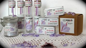 Uma seleção de produtos de açafrão La Casella, incluindo potes, caixas e uma caixa de madeira, dispostos sobre uma superfície texturizada com fios e cristais de açafrão espalhados.