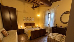 Une chambre confortable avec un lit en bois, une table de chevet avec une lampe, une commode, une chaise et une petite table recouverte d’une nappe à motifs floraux.