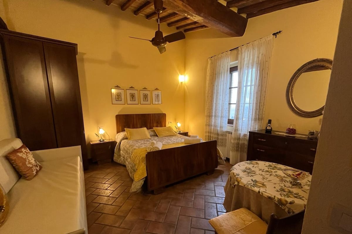 Ein gemütliches Schlafzimmer mit einem hölzernen Bett, einem Nachttisch mit Lampe, einer Kommode, einem Sessel und einem kleinen Tisch mit Blumenmuster.