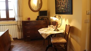 Une pièce comprenant une commode en bois, une table recouverte d’une nappe à motifs floraux, une chaise, un miroir, un tableau et une fenêtre avec des rideaux.