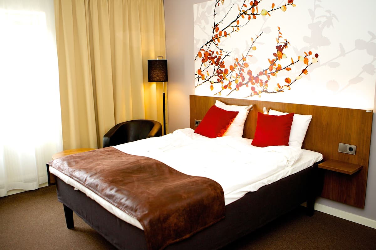 A hotel room with a large bed, a wooden headboard, a red and white pillow, a brown leather throw, a wall-mounted lamp, and a window with curtains.