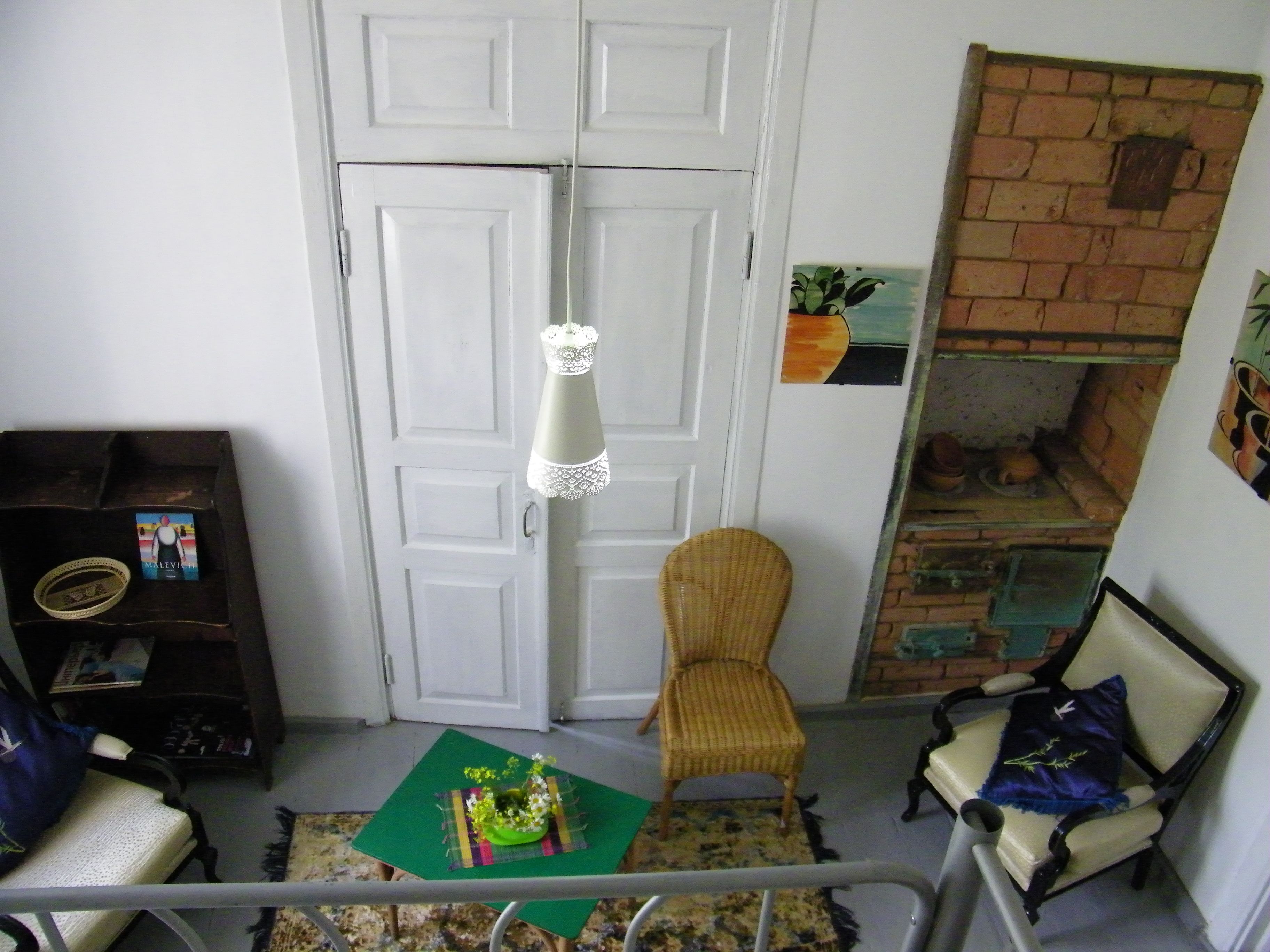 Une pièce avec une porte blanche, une chaise en osier, une petite table avec un set de table vert, une lampe décorative suspendue et un mur en briques agrémenté d’un pot de fleurs.