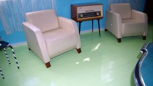 Ein Zimmer mit zwei beigen Sesseln, einem Holztischchen mit einem Radio und einem Streifenmuster-Vorhang.