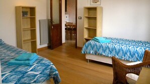 Una habitación con dos camas, suelo de madera, una puerta que da a una cocina y una pequeña silla.