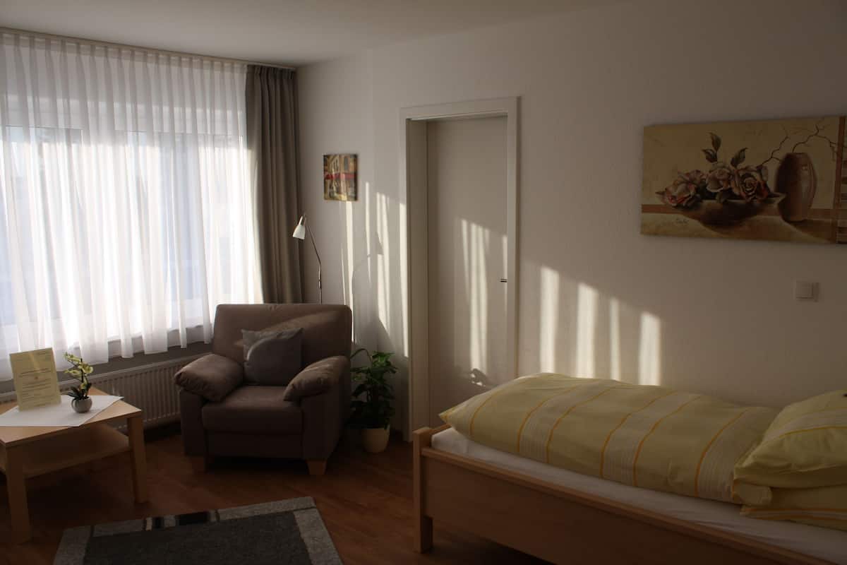 Ein Schlafzimmer mit einem Bett, einem Sessel, einem Schreibtisch, einem Heizkörper und einem Fenster mit durchsichtigen Vorhängen.