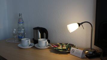 A hotel room with a desk featuring a water bottle, a coffee maker, a cup, a plate with tea bags, a sign, and a remote control.