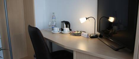 A hotel room with a desk, a chair, a television, a coffee maker, a water bottle, and a cup.