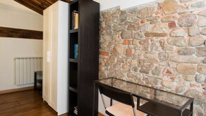 Una habitación con pared de piedra, techo de madera, una estantería y una mesa de cristal con una silla.