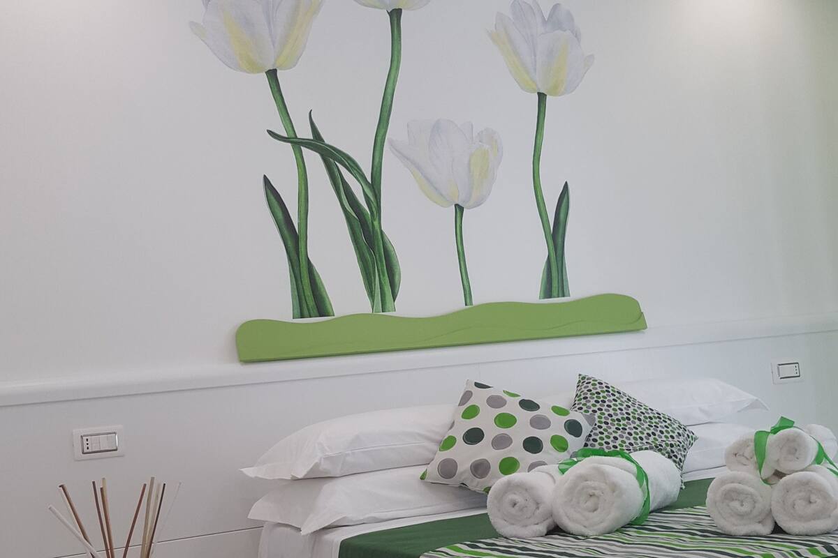 Un dormitorio con cama, almohadas, mesita de noche, un jarrón y un cuadro en la pared.