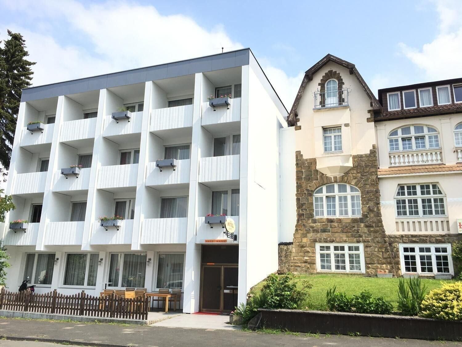 A multi-story hotel with a modern white facade, a stone building with a pitched roof, and a small garden area in front.