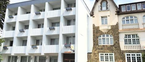 Un hôtel à plusieurs étages, doté d’une façade blanche et moderne, un bâtiment en pierre avec un toit en pente, et un petit jardin à l’avant.