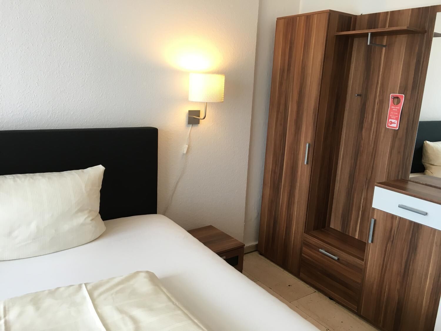 Et hotelværelse med seng, sengebord, garderobeskab og vægmonteret lampe.