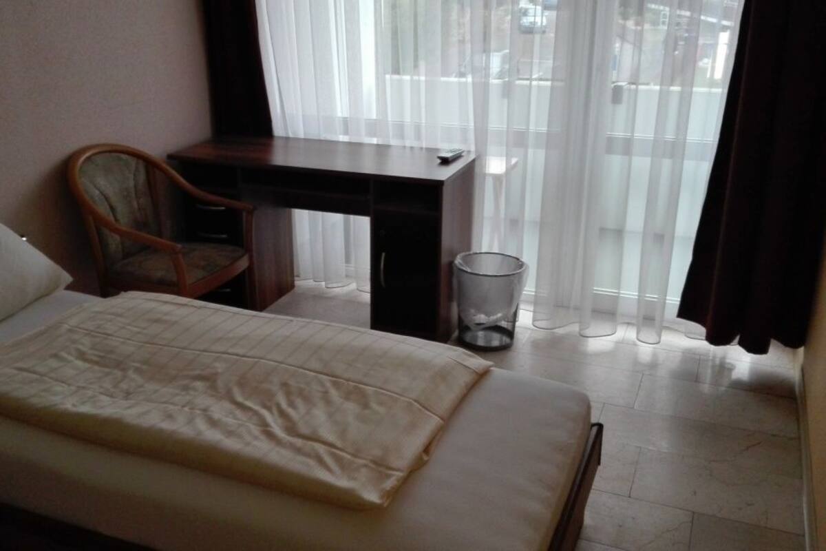 A hotel room with a bed, a chair, a desk, and a window with sheer curtains.