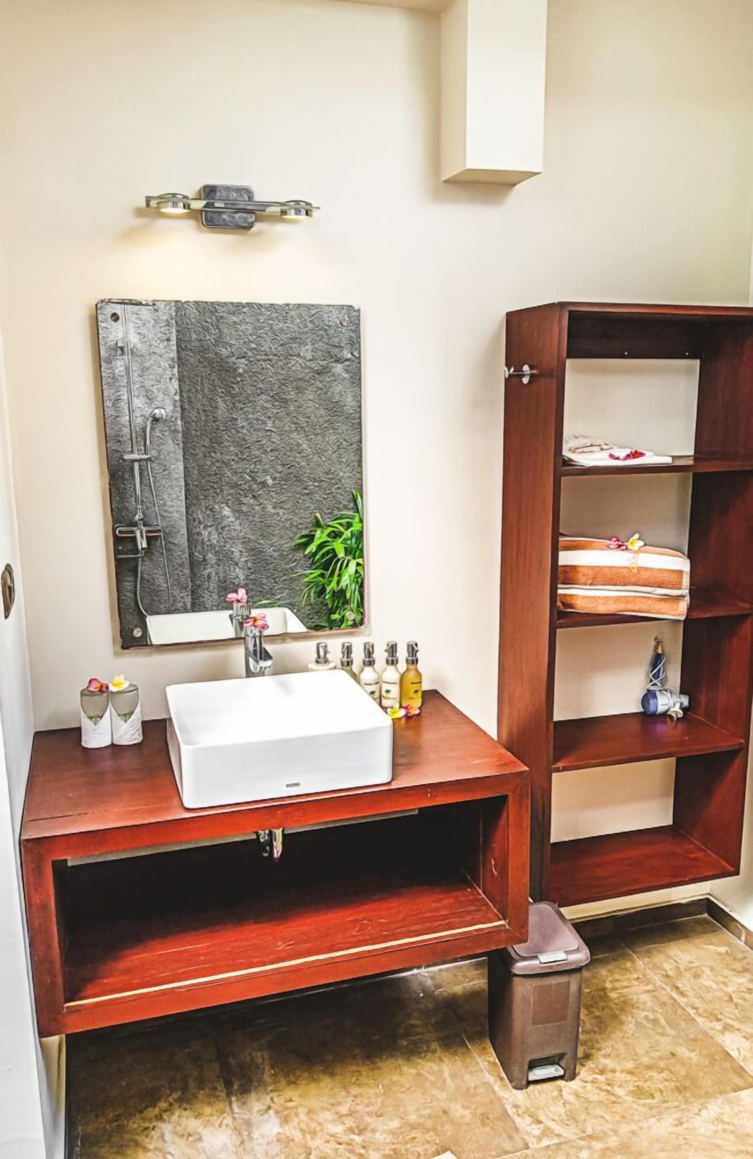 A bathroom with a white sink, a mirror, a shelf with toiletries, and a trash bin.