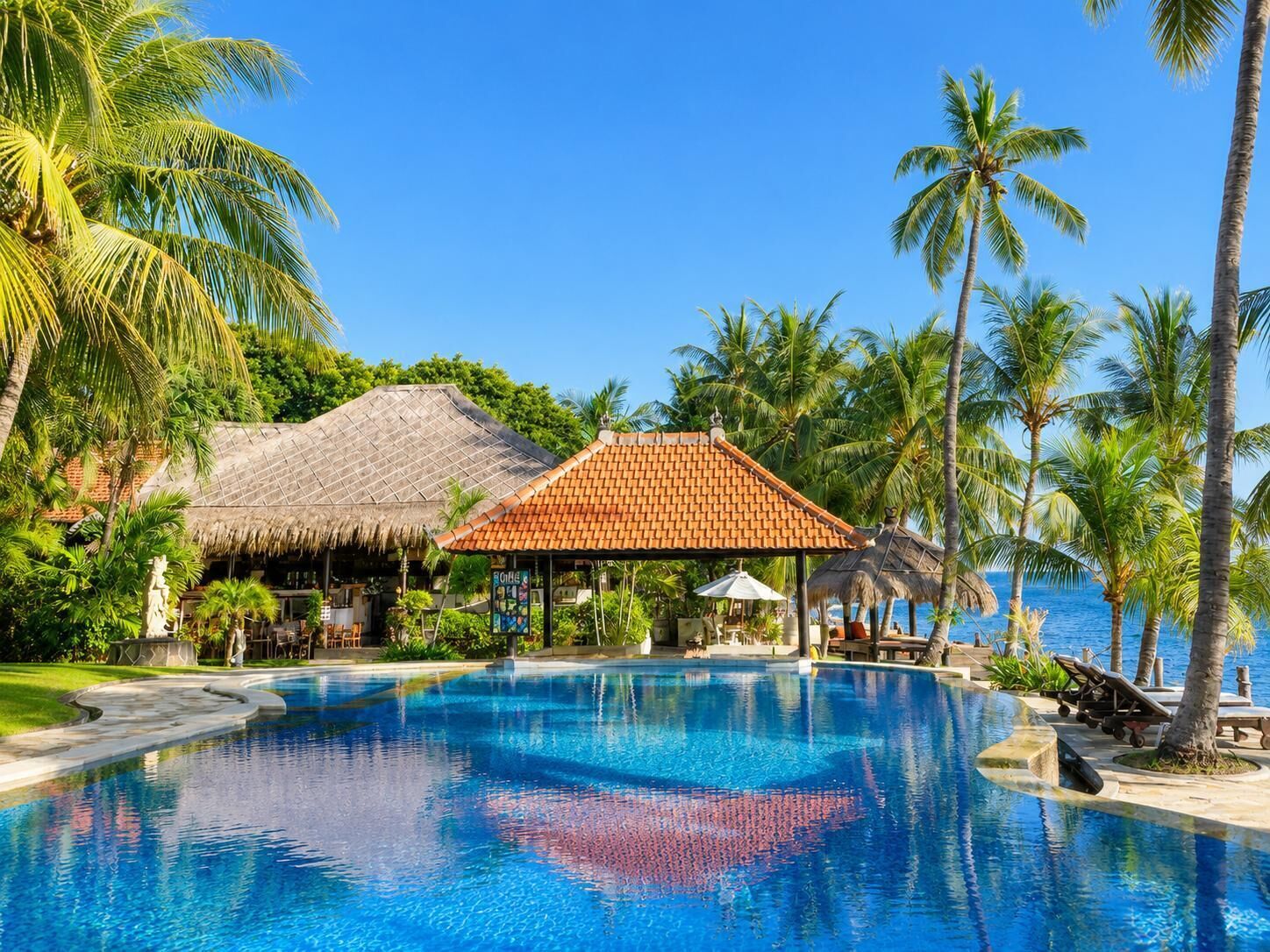 A resort pool with palm trees, a thatched-roof hut, and a clear blue sky.