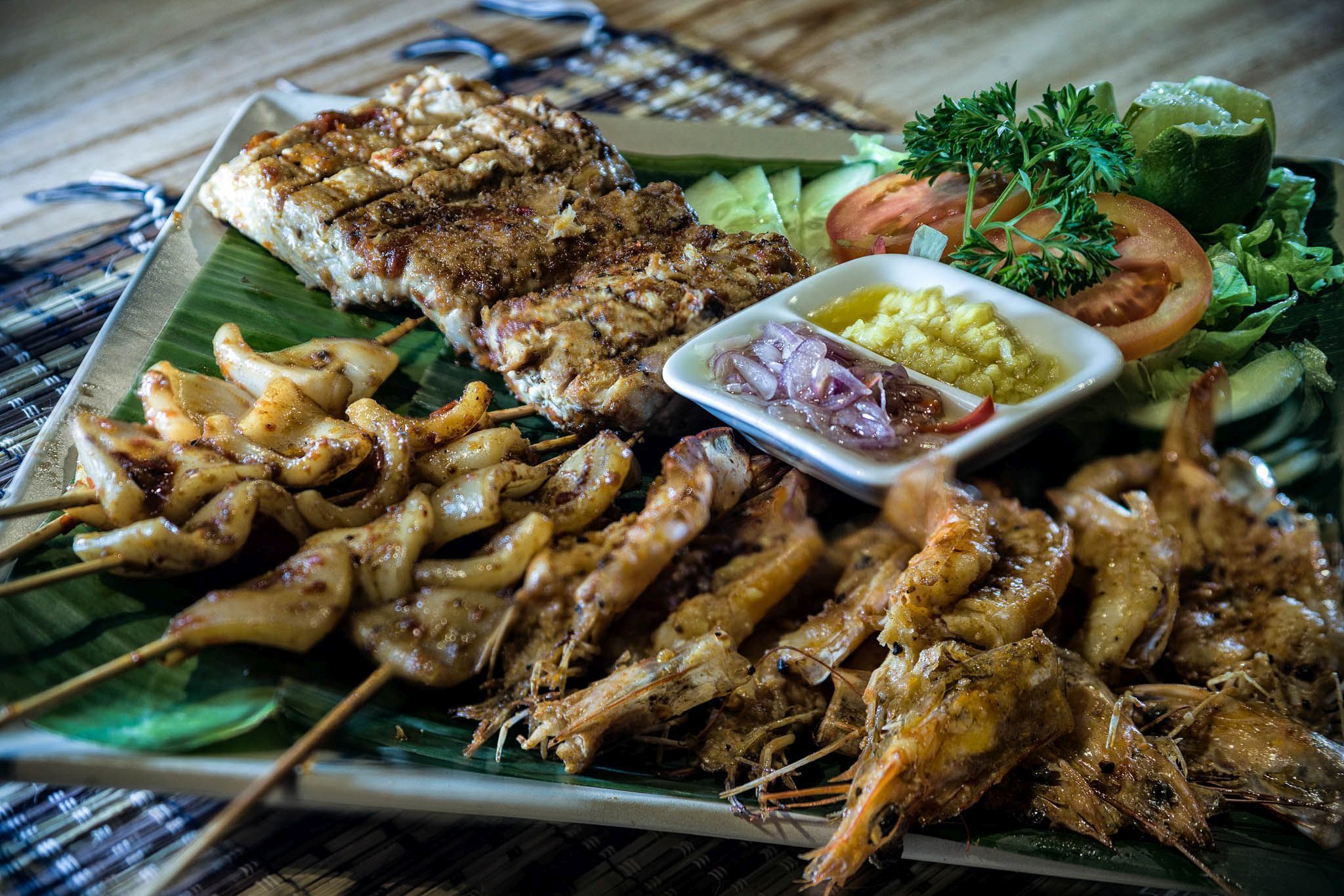Un plato de carnes y mariscos a la parrilla, que incluye brochetas y un bistec, servido sobre una hoja de plátano.