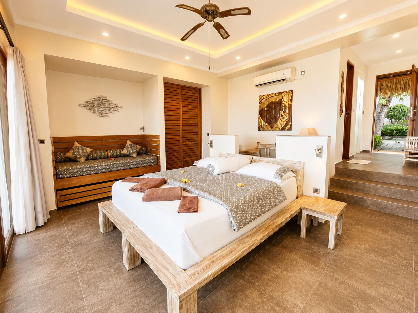 A bedroom with a large bed, a wooden bench, a ceiling fan, and a view of the outdoors.