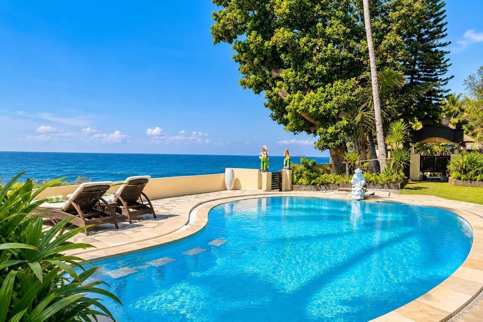 A curved pool with lounge chairs and a statue, overlooking a clear blue ocean.