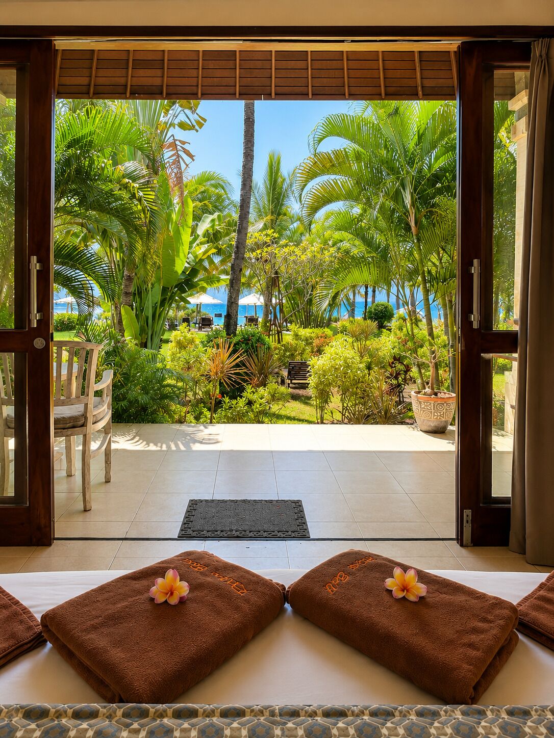 A room with a view of a tropical garden and the sea, featuring a bed with towels and a potted plant.