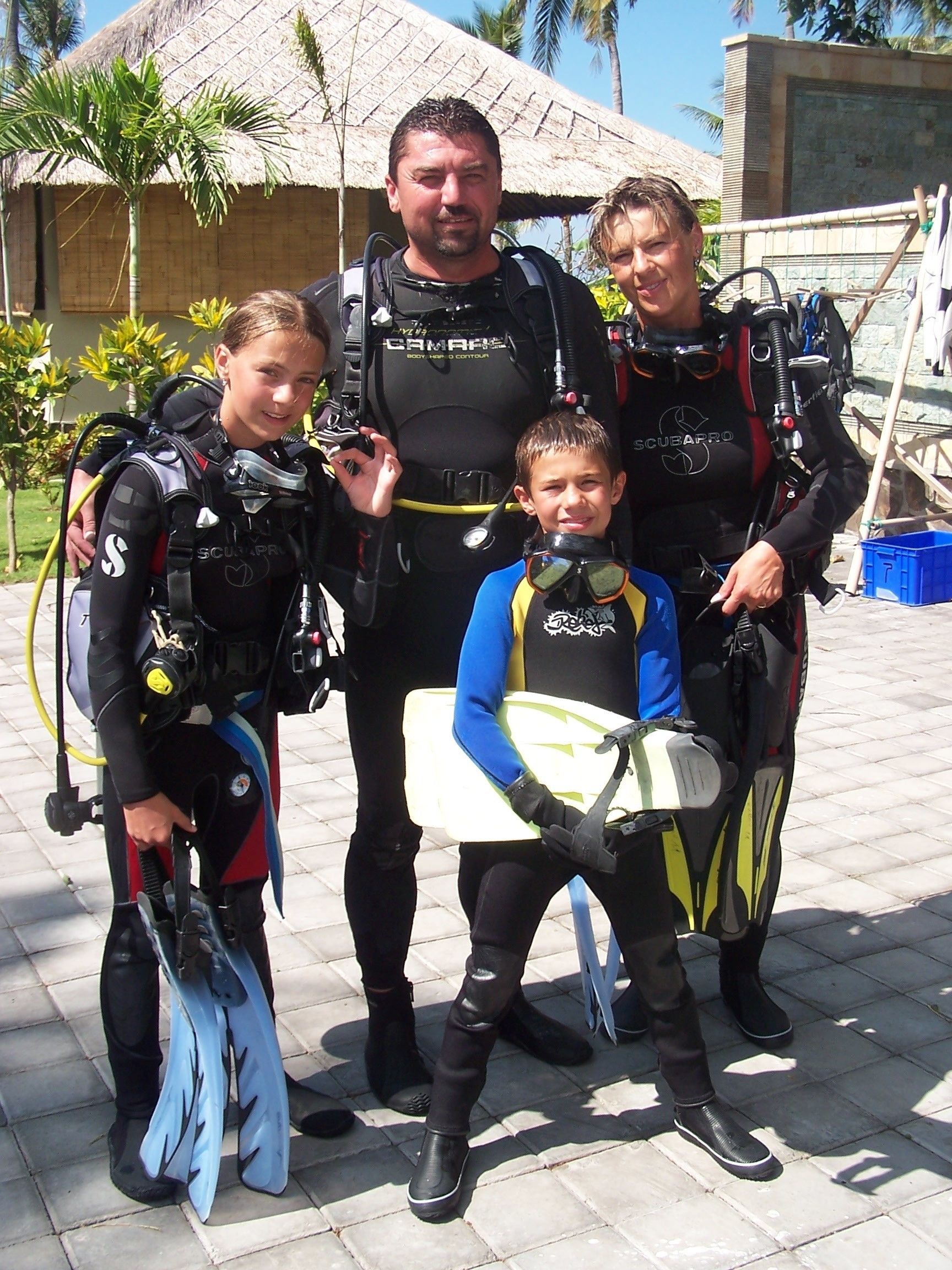 A family with scuba diving gear, including wetsuits and fins, standing in front of a building with a thatched roof and palm trees.