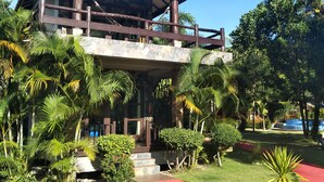 Un edificio de dos pisos con techo rojo y balcón de madera, rodeado de abundante vegetación y palmeras.