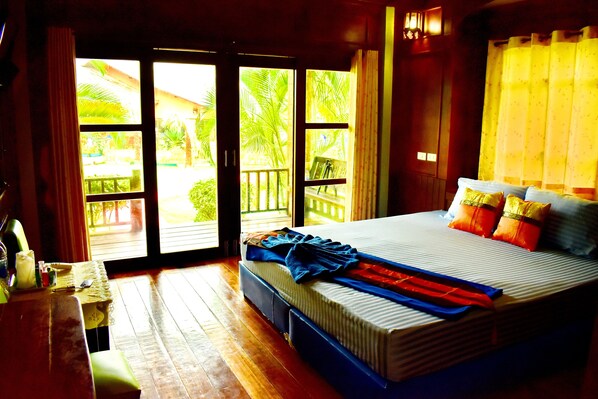 A bedroom with a large bed, wooden floor, and a view of palm trees through the windows.