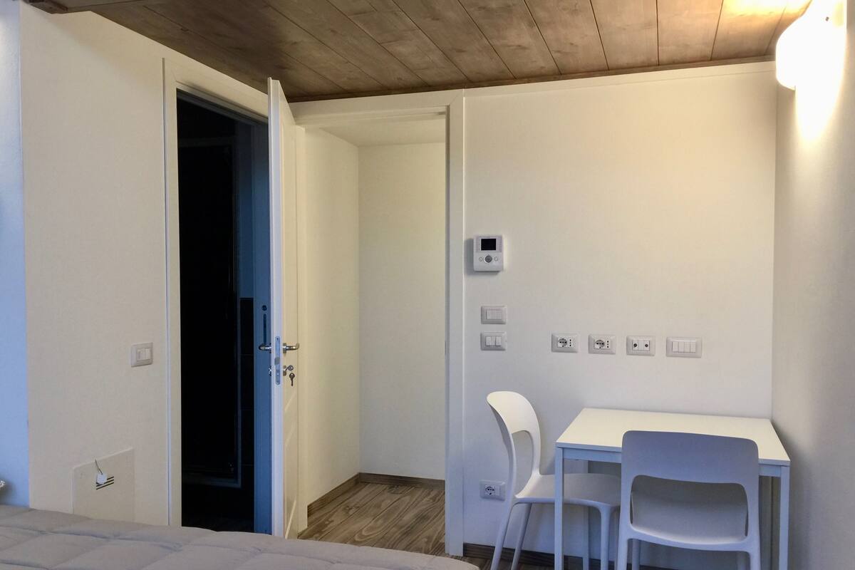 Un dormitorio con cama, una puerta abierta que da al baño, un escritorio pequeño con silla e un termostato en la pared.