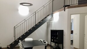 A dining area with a black table and white chairs, a black bookshelf, and a staircase with a black railing. - Zannetti (Florence)