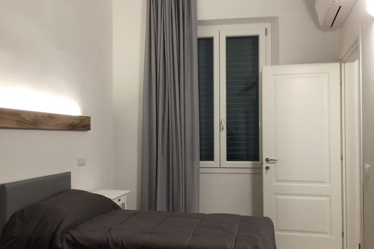 Un dormitorio con cama, mesita de noche, ventana con cortinas y una puerta.
