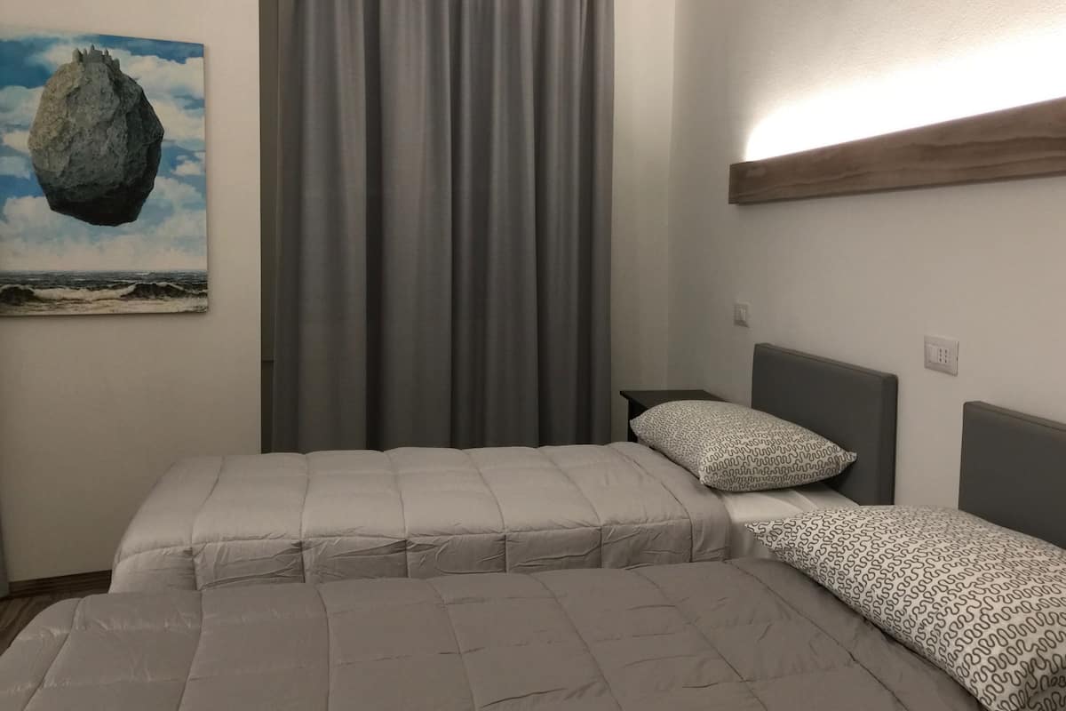 Un dormitorio con dos camas, cortinas grises, un cuadro de una roca y una mesita de noche con un cojín estampado.