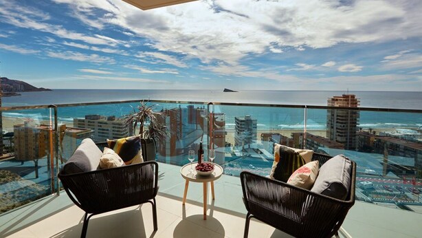 A balcony with two wicker chairs, a small round table with a bowl of fruit and a bottle, and a glass-top table with two wine glasses. The view includes a beach and buildings across the water. - Sunset Drive Holidays Benidorm (Benidorm)