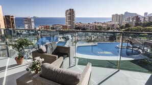 A rooftop terrace with a glass balustrade, wicker furniture, a potted plant, and a view of a swimming pool and cityscape. - Sunset Drive Holidays Benidorm (Benidorm)