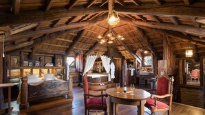 A rustic wooden interior with a high ceiling, a round wooden table with chairs, a bed with a canopy, and a fireplace.