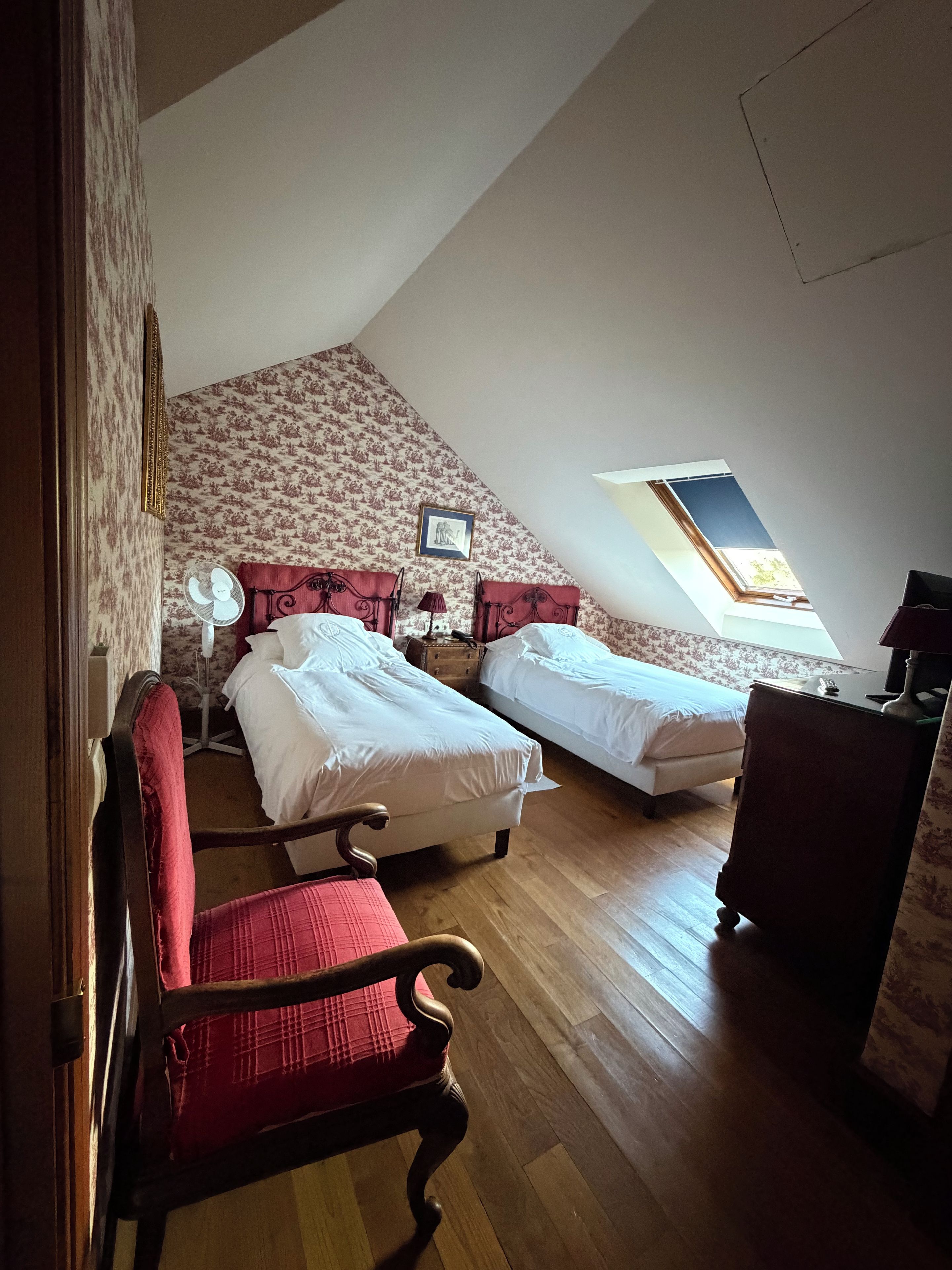 Een kamer met twee bedden, een houten vloer, een rode stoel, een bureau en een raam met uitzicht.