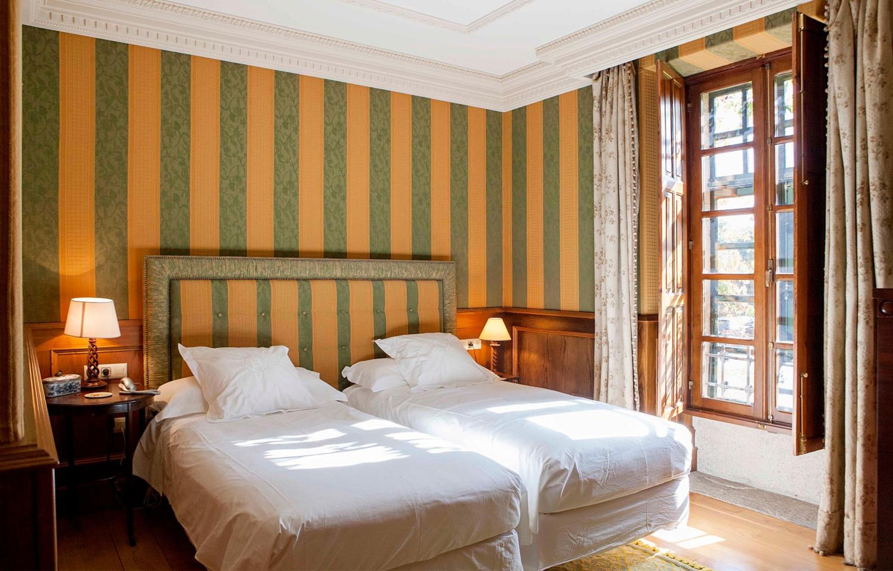 A hotel room with two beds, striped wallpaper, a wooden headboard, a nightstand with a lamp, and a window with curtains.