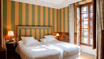 A hotel room with two beds, striped wallpaper, a wooden headboard, a nightstand with a lamp, and a window with curtains.
