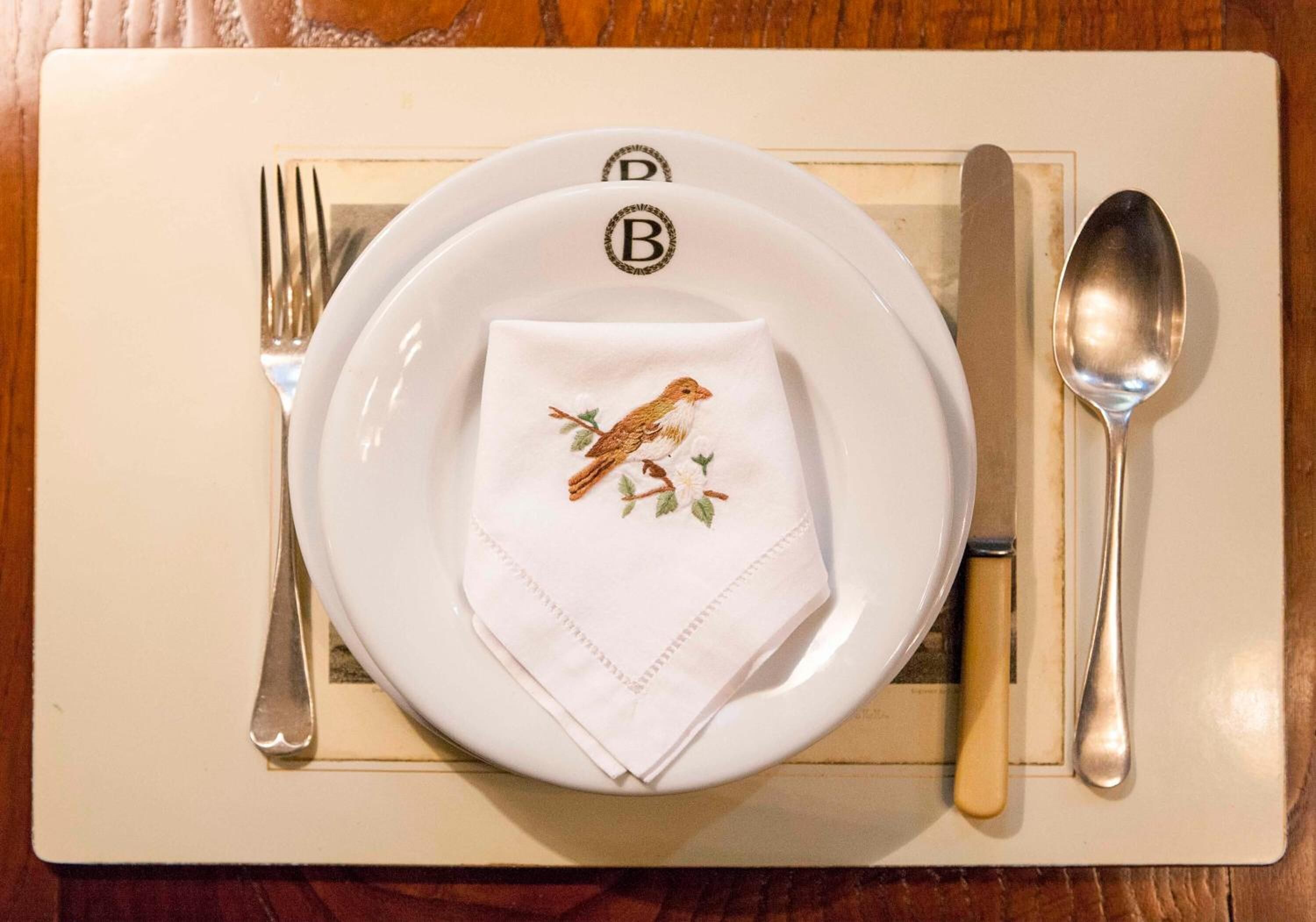 A neatly set dining table with a plate, a folded napkin with a bird embroidery, a fork, a knife, and a spoon.