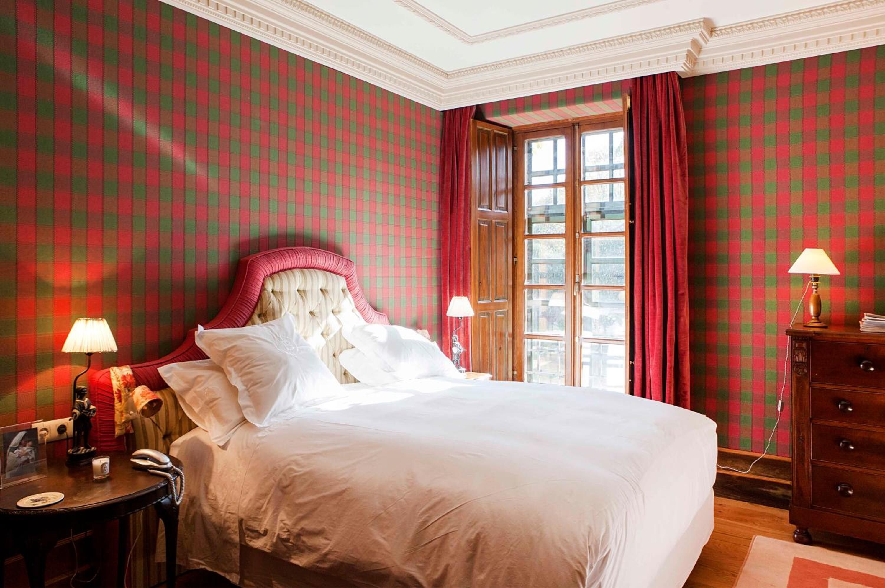 A hotel room with a checkered wallpaper, a large bed with white linens, two bedside lamps, a wooden dresser, and a window with red curtains.
